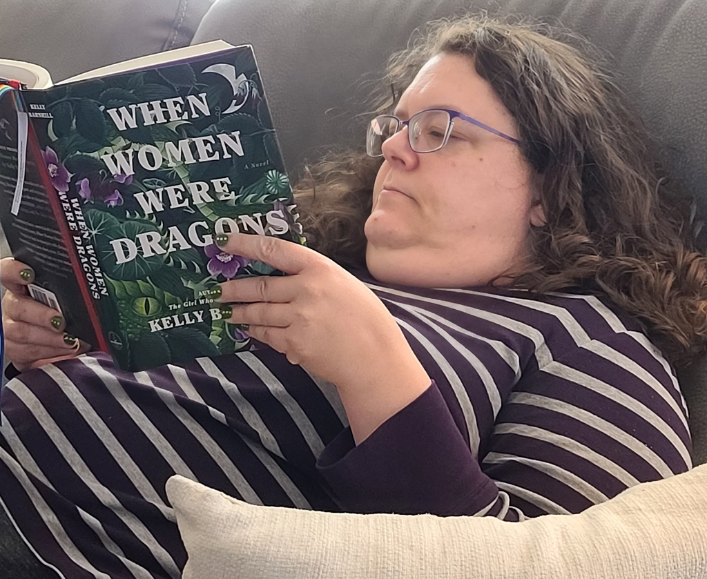 Photo of Ms. Pointer wearing a purple and gray striped shirt laying on her couch reading the book When Women Were Dragons by Kelly Barnhill. 